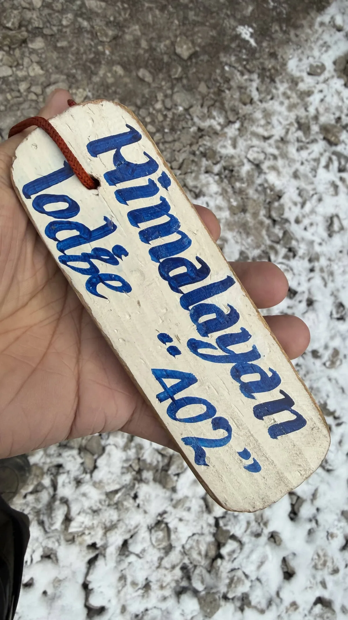 手持一塊木製房間鑰匙牌,藍字寫著 Himalayan Lodge 402,地上積雪